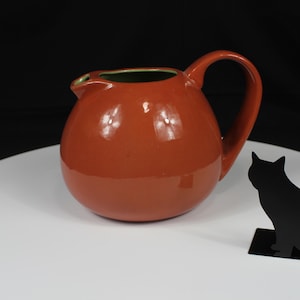 May include: A spherical, orange ceramic jug with a handle and a green interior rim. A black cat silhouette is next to the jug on a white surface. The background is black.