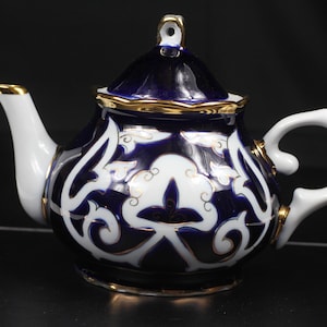 Könnte beinhalten: Eine Keramik-Teekanne mit einem marineblauen und weißen Blumenmuster. Die Teekanne hat einen weißen Griff und Ausguss mit goldenen Zierleisten. Der Deckel ist marineblau mit goldenen Zierleisten und einem kleinen Schlaufengriff. Auf schwarzem Untergrund.