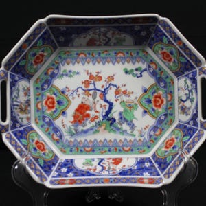 May include: An octagonal ceramic serving dish with two integrated handles, featuring a white base decorated with colorful hand-painted scenes of figures under a tree, surrounded by intricate blue, green, and orange floral and geometric patterns.