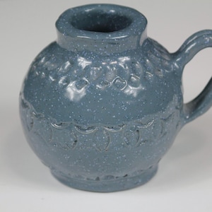 May include: A small, round, blue ceramic pitcher with a speckled finish and a single handle. The body of the pitcher features two horizontal bands of embossed, scalloped, and circular patterns encircling the exterior surface of the vessel.