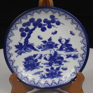 May include: A white ceramic plate with a blue border and a detailed blue illustration. The illustration depicts a tree, figures, butterflies, and floral elements. The plate is displayed on a wooden stand.