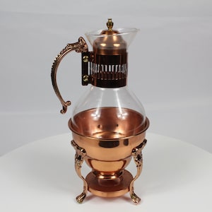 May include: A clear glass carafe with a copper-toned metal frame, handle, and base. The carafe features a decorative metal collar with vertical slats, a matching copper lid with a small finial, and a stand supported by three ornate legs.