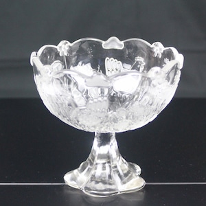 Puede incluir: Un cuenco de cristal transparente sobre pedestal con borde festoneado y detalles en relieve de gallos y motivos florales. El cuenco tiene un recipiente ancho y redondeado sostenido por una base robusta y acampanada, con acabado transparente.