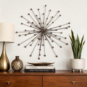 May include: A starburst wall sculpture made of thin dark metal rods with small spheres at the ends. The sculpture is mounted on a wall above a wooden console table that holds a lamp, a speckled vase, a decorative bowl, and two stacked books.
