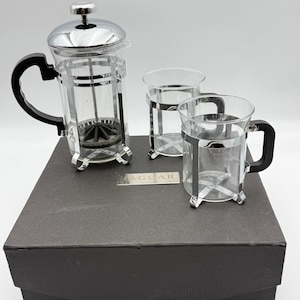 May include: A glass French press coffee maker with a chrome lid and black handle, accompanied by two matching glass mugs with chrome frames and black handles. A small rectangular metal plaque with the text JAGLAR is visible.