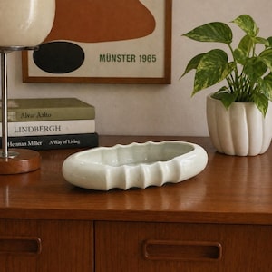 May include: A cream-colored, oblong ceramic bowl with a ridged design. The bowl sits on a wooden surface, alongside a potted plant and a stack of books. A framed print with the text "MÜNSTER 1965" hangs in the background.