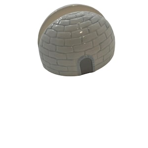 May include: A ceramic napkin holder shaped like an igloo. The igloo is light gray with a brick pattern and a small arched doorway. A curved, off-white section forms the top of the holder, designed to hold napkins. The holder is on a white background.