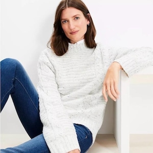 May include: A light gray cable knit sweater with flecks of color. The sweater has a mock neck and long sleeves. The person in the image is wearing blue jeans. The sweater is a cozy and stylish garment.