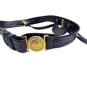 Pode incluir: Um cinto de couro preto texturizado com uma fivela circular de metal dourado apresentando um emblema de águia em relevo. O cinto inclui várias tiras, passadores de metal na cor latão e uma pequena aba de couro com rebites metálicos.