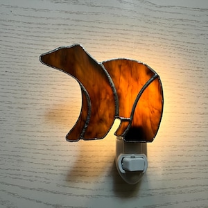 May include: A stained glass night light in the shape of a bear. The bear is made of amber and brown glass, with a silver-colored metal outline. The night light has a white plastic base with an on/off switch.