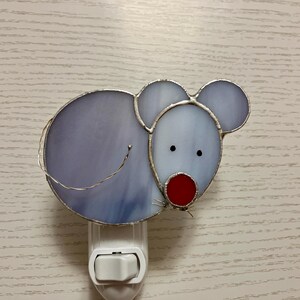 May include: A stained glass nightlight shaped like a mouse with a purple body, a red nose, and black eyes. The nightlight is plugged into a white electrical outlet.