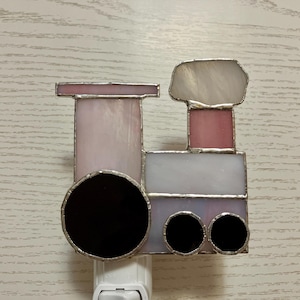 May include: A stained glass night light shaped like a train engine. The train is pink, white, and black. The night light has a standard plug-in base.