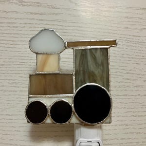 May include: A stained glass night light shaped like a train with a white smoke stack, brown and green glass, and black wheels.