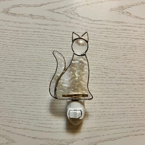 May include: A decorative night light shaped like a cat. The cat is made of clear, textured glass with a silver-colored outline. The night light plugs into a standard wall outlet and has an on/off switch.