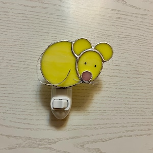 May include: A yellow stained glass night light in the shape of a mouse. The mouse has black eyes, a pink nose, and silver whiskers. The night light is plugged into a white outlet.