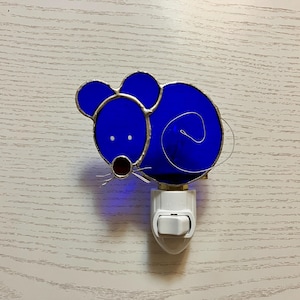 May include: A blue stained glass night light in the shape of a mouse. The mouse has two small white eyes, a red nose, and wire whiskers. The night light is plugged into a white outlet.