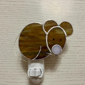 May include: A stained glass nightlight shaped like a brown mouse with a white nose and black eyes. The nightlight is plugged into a white electrical outlet.