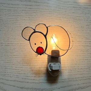 May include: A stained glass night light shaped like a mouse. The mouse has a red nose, black eyes, and wire whiskers. The light bulb is lit, and the night light is plugged into a wall outlet.
