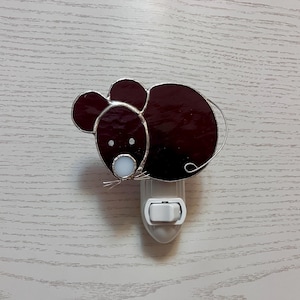May include: A stained glass night light in the shape of a mouse. The mouse has a dark burgundy body and ears, with a white face and small white whiskers. The night light is plugged into a white outlet.