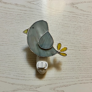 May include: A stained glass night light shaped like a bird. The bird has a gray body, yellow beak and feet, and a small black eye. The night light plugs into a wall outlet and has an on/off switch.