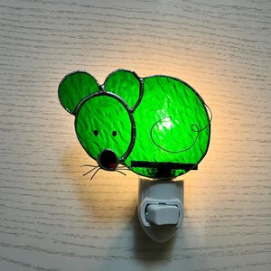 May include: A green stained glass night light in the shape of a mouse. The mouse has large ears, a round body, and a black nose. The night light plugs into a standard wall outlet and has an on/off switch.