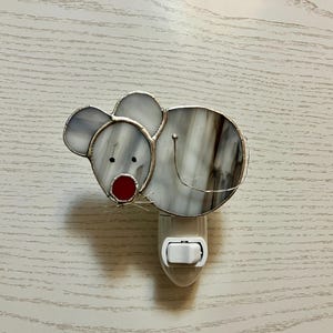 May include: A stained glass night light in the shape of a mouse. The mouse has gray and white glass with a red nose and black eyes. The night light has a white on/off switch and is plugged into an outlet.