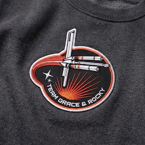 Op de afbeelding: Donkergrijze sweatshirt met een cirkelvormige patch met een raketontwerp en de tekst "TEAM GRACE & ROCKY". De patch heeft een zwarte achtergrond met oranje en witte accenten en een sterrenafbeelding.