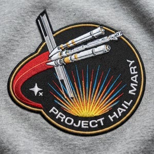 May include: Embroidered patch featuring a spaceship design with the text "PROJECT HAIL MARY." The patch has a black background with a gold border, a red and white curved accent, and a star. The spaceship is white and gold.