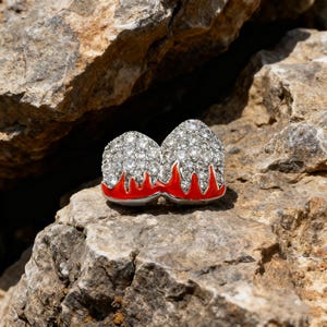 May include: A distinctive ring with two gem-covered domes, set above a red enamel base. The ring is displayed on a natural stone surface, showcasing its sparkling appearance. The design is striking and unusual.