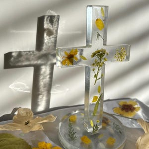 Peut inclure: Une croix en résine transparente avec des fleurs jaunes et de la verdure incrustées. La croix est montée sur une base ronde transparente, contenant également des fleurs séchées. L'ombre de la croix est visible sur le fond blanc.