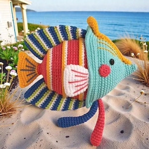 Könnte beinhalten: Ein farbenfrohes, gehäkeltes Fischspielzeug mit einem skurrilen Design. Der Fisch hat einen türkisfarbenen Kopf, einen roten und orangefarbenen Körper und gestreifte Flossen in Blau, Grün und Gelb. Er hat eine weiße Flosse, eine rote Wange und einen hängenden roten Schwanz. Das Spielzeug liegt am Sandstrand.