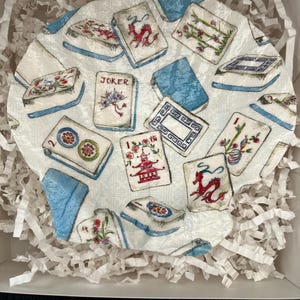 May include: A round, white decorative plate featuring a pattern of Mahjong tiles with red, blue, and green accents. The plate is surrounded by blue fabric and white paper filler, presented in a white box.