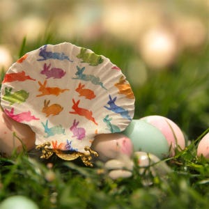 May include: A shell-shaped Easter decoration featuring colourful bunny illustrations. The decoration sits on a gold stand, surrounded by green grass and pastel Easter eggs. The bunnies are in shades of pink, blue, and orange, creating a festive scene.