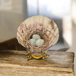 Puede incluir: Concha decorativa con una escena de nido pintada. La concha presenta un nido con tres huevos moteados, rodeado de flores rosas y una pluma. La concha está sostenida por un soporte de metal dorado.