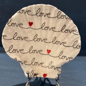 May include: A white seashell decorated with the word "love" in black script. The word is repeated across the shell, interspersed with small red glitter hearts. The shell is displayed on a silver metal stand.