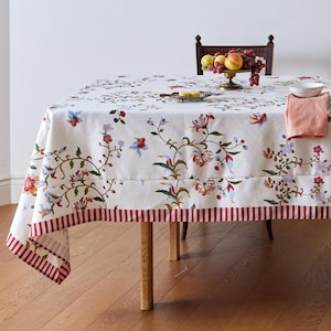 May include: A rectangular tablecloth featuring a white base with a colorful floral and vine pattern in shades of red, blue, and green. The bottom edge is finished with a wide border of red and white vertical stripes, creating a distinct contrast.