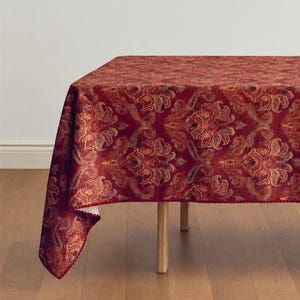 May include: A rectangular tablecloth featuring a deep red base color with an intricate, repeating damask and paisley pattern in shades of gold and copper. The fabric has a textured appearance and is draped over a table with visible wooden legs.