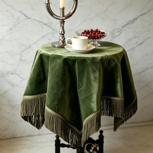 May include: A small round table draped with a green velvet tablecloth featuring a fringe trim. A white teacup and saucer sit on the table, along with a glass bowl of cherries and a silver candelabra.