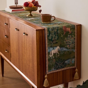 May include: A wooden sideboard with a decorative tapestry runner featuring a forest scene with a white horse and a lion. A bronze bowl of fruit and a brown mug sit on top. The runner has gold tassels.