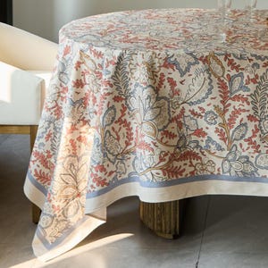 Può includere: Una tovaglia rotonda con base color crema caratterizzata da un motivo floreale e botanico ripetuto in tonalità di rosso spento, blu polvere e giallo tenue. Il tessuto presenta un ampio bordo lungo il perimetro con una striscia blu.