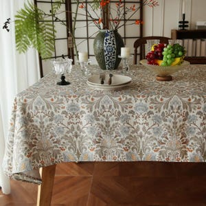 Puede incluir: Un mantel rectangular con un patrón botánico repetitivo en tonos apagados de verde salvia, azul polvoriento y naranja terracota sobre un fondo crema, colocado sobre una mesa de madera con un jarrón de cerámica, frutero y cristalería.