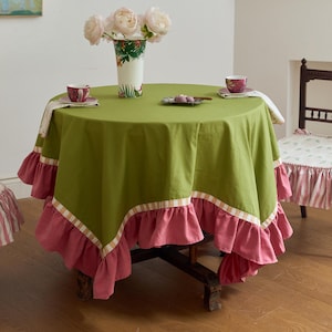 May include: A round table covered with a solid olive green tablecloth featuring a wide, ruffled pink border and a chequered trim. The table is set with a floral vase containing white peonies, two patterned teacups, and a small rectangular plate.