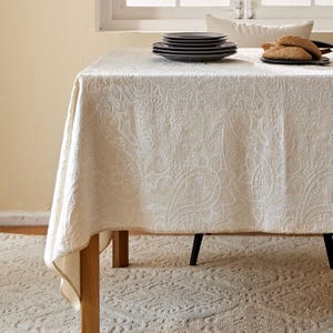 Op de afbeelding: Crèmekleurig paisley tafelkleed op een houten tafel. Het tafelkleed heeft een ingewikkeld, reliëf ontwerp. Zwarte borden en brood liggen op de tafel. De tafel staat op een patroonkleed.