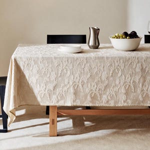 May include: A cream-coloured rectangular tablecloth featuring a raised, textured floral pattern across its surface. The fabric drapes over a wooden table, which is set with a stack of white plates, a metallic pitcher, and a bowl of fruit.