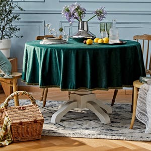 May include: A round table covered with a dark green velvet tablecloth featuring a decorative trim along the edge. The table is set with a silver vase containing purple flowers, a glass carafe, and a plate of yellow lemons on a silver tray.