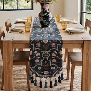 May include: A dark blue table runner featuring a repeating floral and botanical pattern in shades of pink, cream, and green. The runner is finished with a decorative border and a series of hanging blue tassels along the pointed bottom edge.