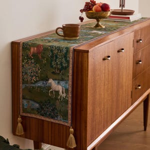 May include: A wooden sideboard with a green tapestry runner featuring a lion and a white horse in a forest scene. A brown mug and a brass bowl of fruit sit on top. The runner has gold tassels.