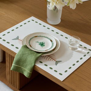 May include: A rectangular white placemat featuring a green leaf border and two fish skeleton illustrations. A stack of two white ceramic plates with green floral and geometric patterns sits on the mat, topped with a folded olive green linen napkin.
