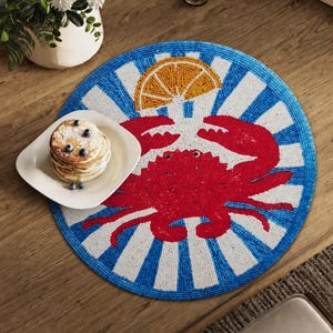 May include: A round beaded placemat featuring a vibrant red crab centered against a background of alternating blue and white rays, with a yellow citrus slice motif above the crab. A square white plate holds a stack of pancakes topped with blueberries.
