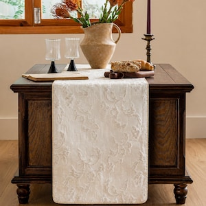 Puede incluir: Un camino de mesa de color crema con un patrón floral de damasco en relieve tono sobre tono y un brillo sutil. El camino rectangular tiene bordes acabados y está colocado sobre una mesa de madera marrón oscuro, sirviendo de base para una jarra.
